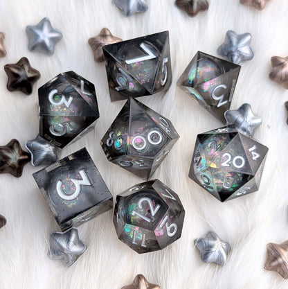 A set of translucent smoky gray polyhedral gaming dice with holographic glitter and white numbering, arranged on a white faux fur surface with scattered metallic star-shaped props.