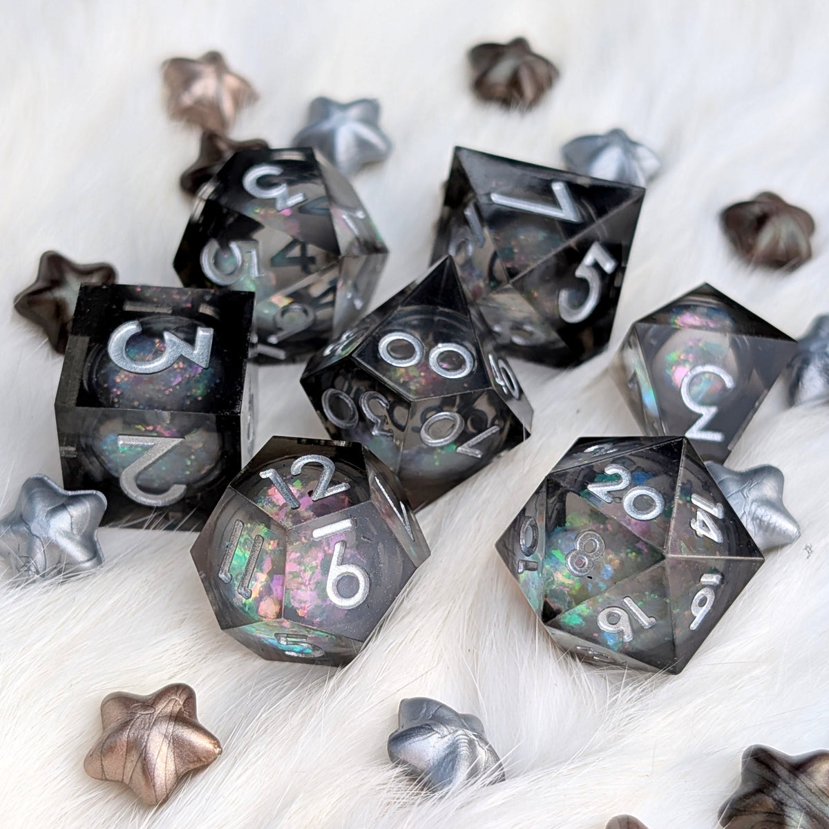 A set of translucent black polyhedral gaming dice with holographic glitter and white numbering, arranged on a white faux fur surface with decorative metallic star-shaped props.