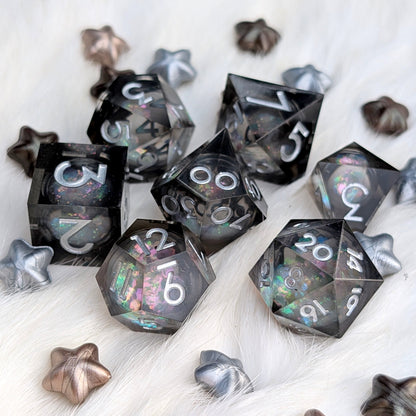 A set of translucent black polyhedral gaming dice with holographic glitter and white numbering, arranged on a white faux fur surface with decorative metallic star-shaped props.