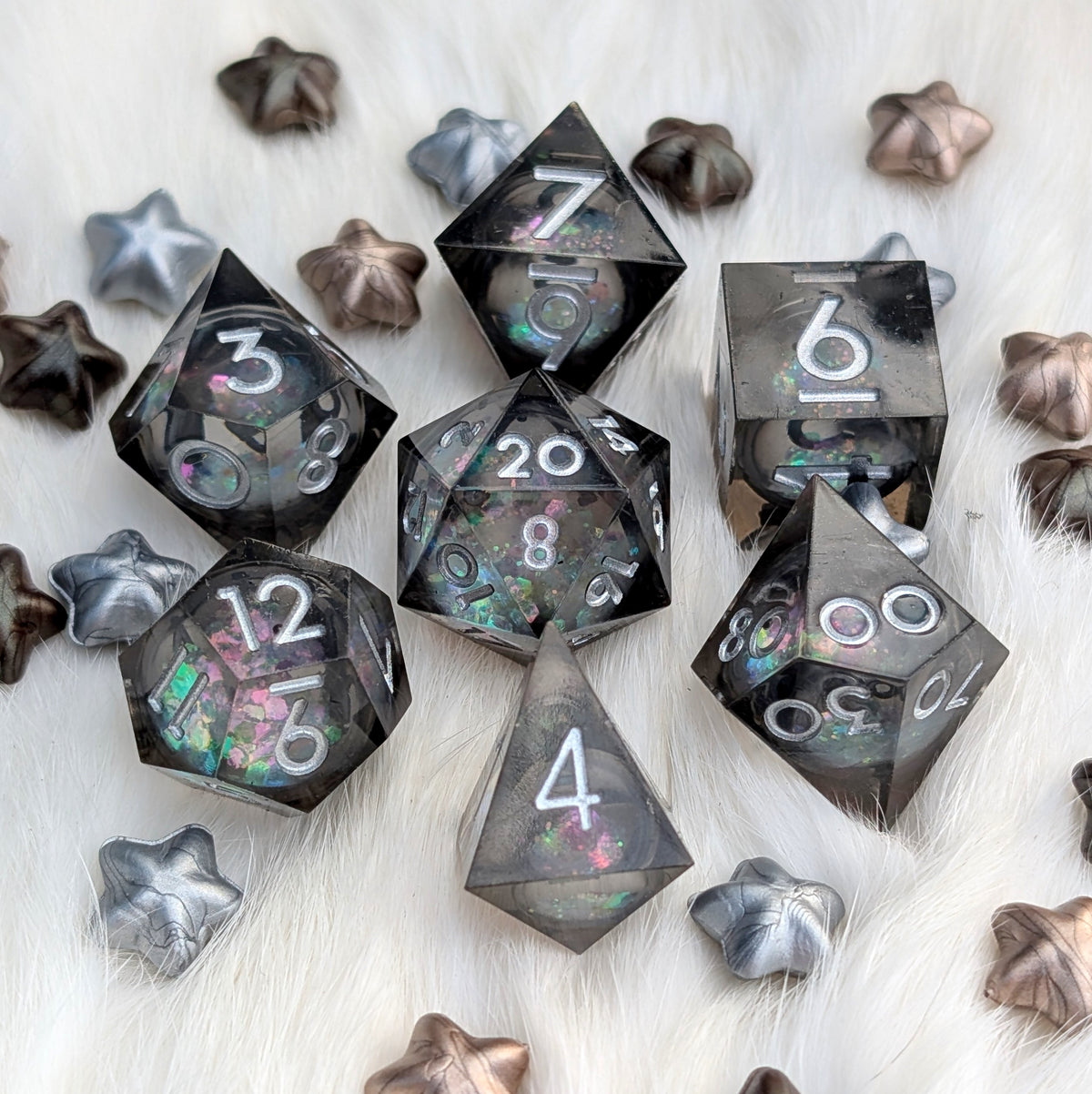 A set of translucent smoky gray polyhedral dice with iridescent glitter and white numbering, arranged on a white faux-fur surface with metallic star-shaped props.