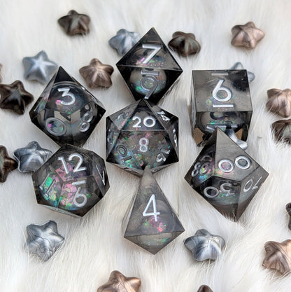 A set of translucent smoky gray polyhedral dice with iridescent glitter and white numbering, arranged on a white faux-fur surface with metallic star-shaped props.