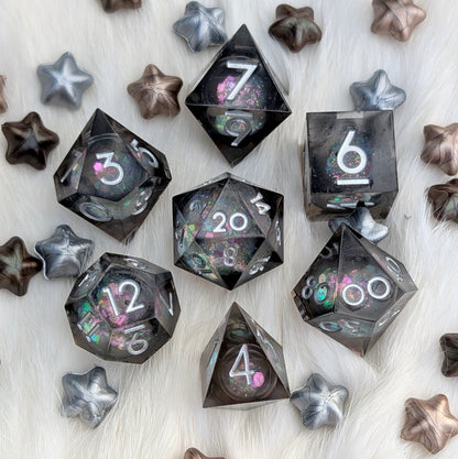 A set of translucent dark gray polyhedral gaming dice with holographic glitter and white numbering, arranged on a white faux fur surface with scattered metallic star-shaped props.