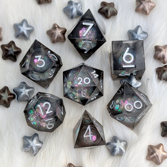 A set of translucent dark gray polyhedral gaming dice with holographic glitter and white numbering, arranged on a white faux fur surface with scattered metallic star-shaped props.