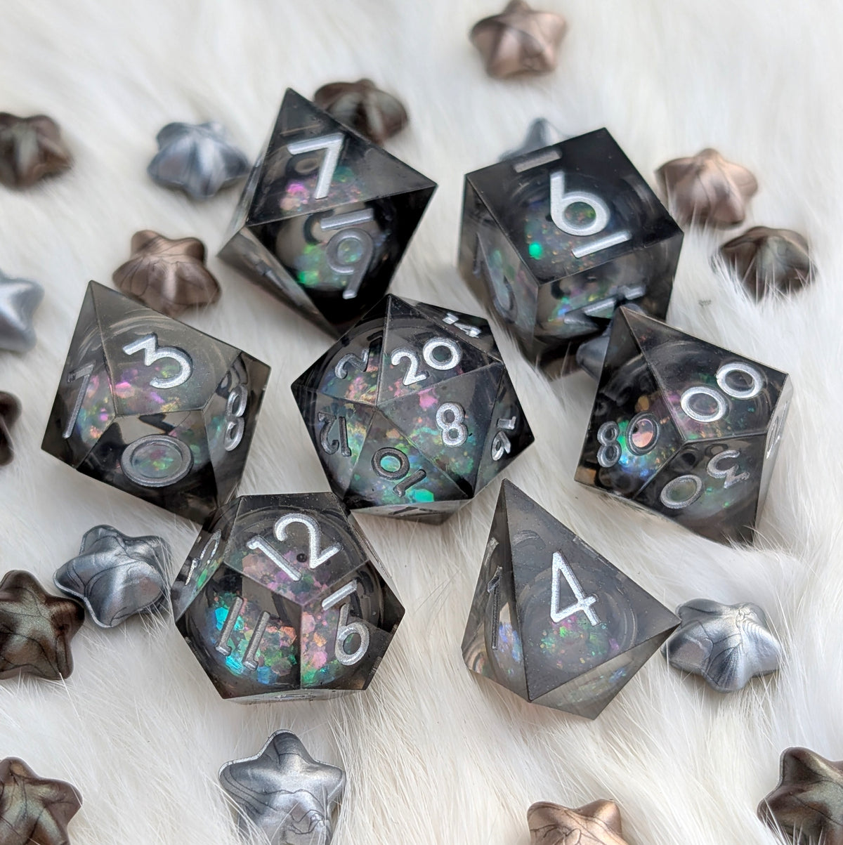 A set of translucent smoky gray polyhedral gaming dice with iridescent glitter and white numbering, arranged on a white faux-fur surface with matching metallic star-shaped props.