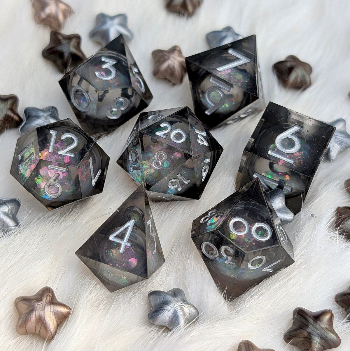 A set of translucent smoky black polyhedral gaming dice with holographic glitter and white numbering, arranged on a white faux fur surface with decorative metallic star-shaped props.