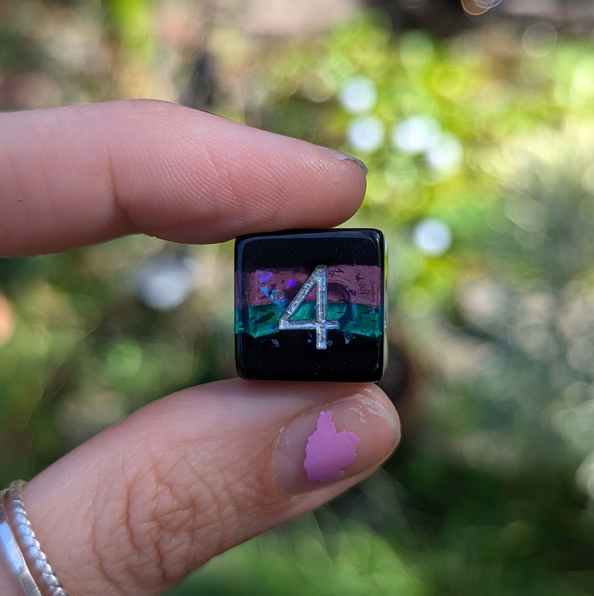 Hand holding a black and multicolored die with glitter, number four visible, part of the Midnight Nebula Dice Set against a blurred background.