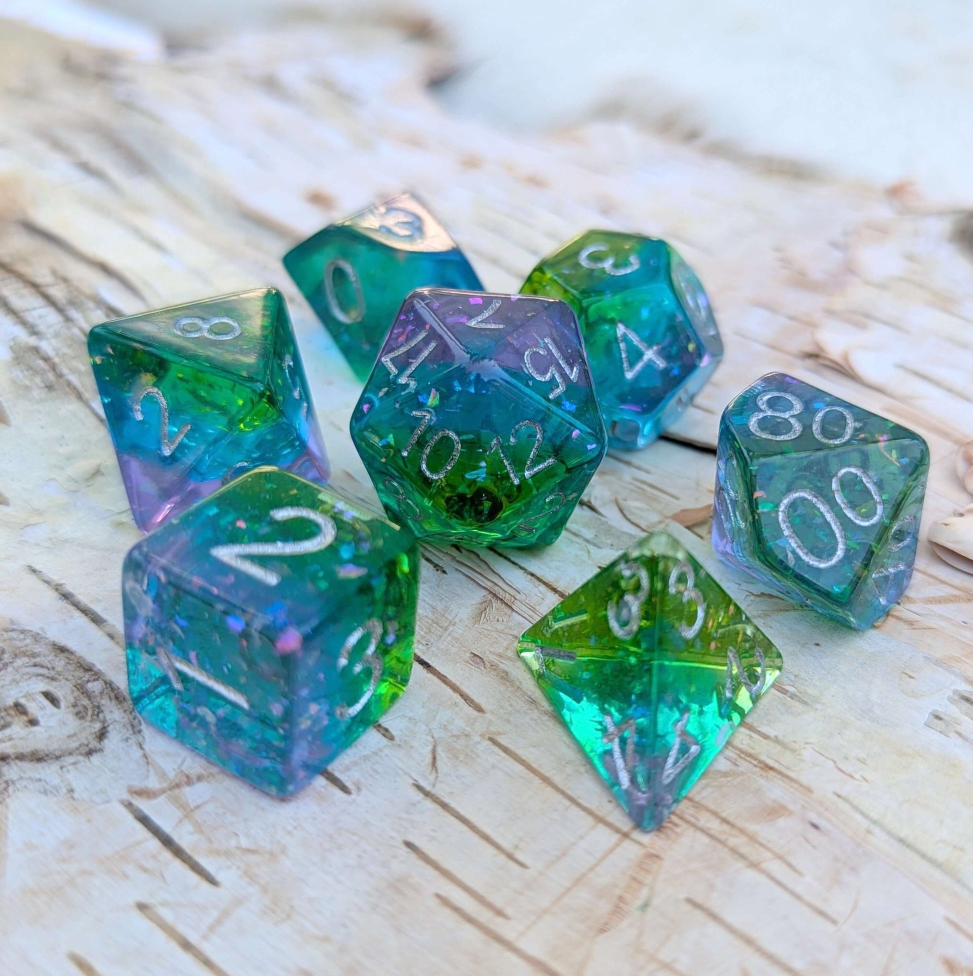 Aurora Sparkle Dice Set with green, blue, and purple hues, iridescent glitter, on a wood surface. Perfect for celestial-themed adventures.