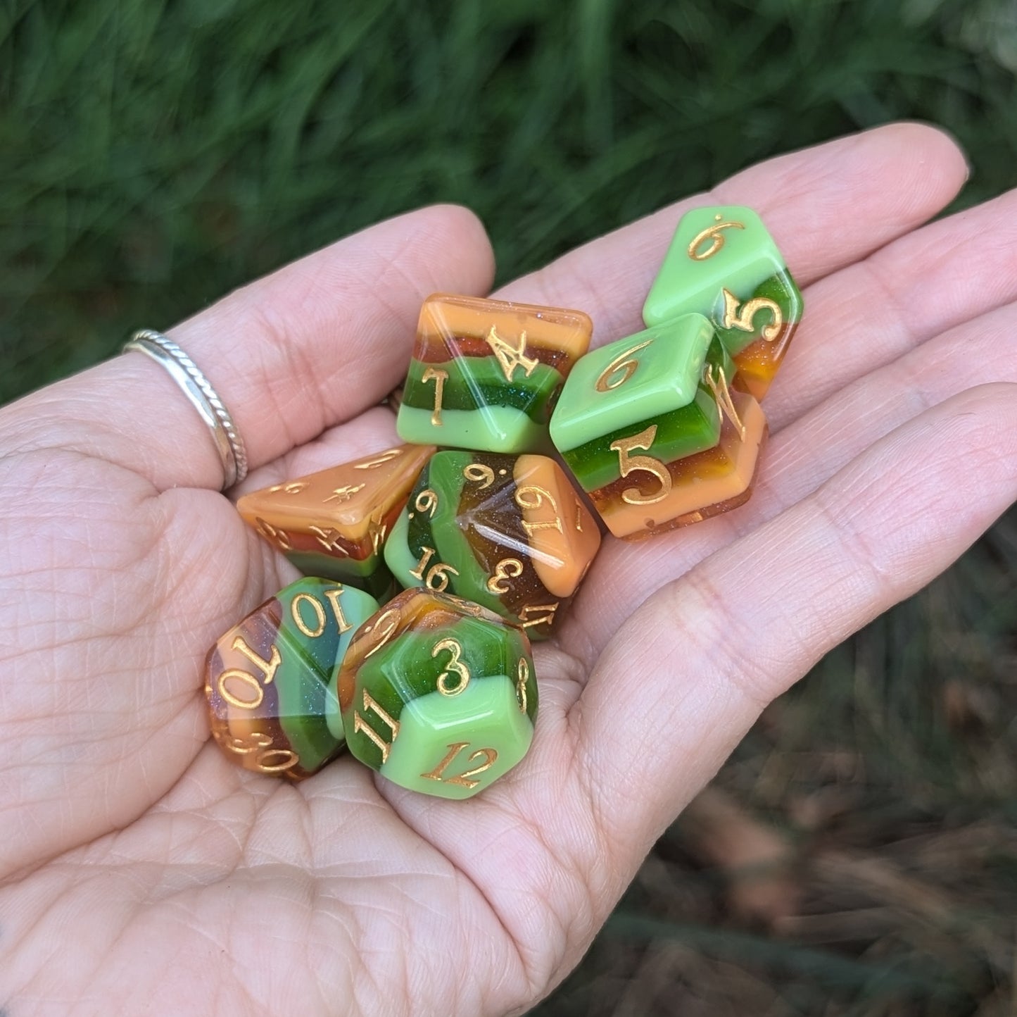 Hand holding Timberlight Dice Set with ombre greens, browns, and gold font, against a background of natural grass.