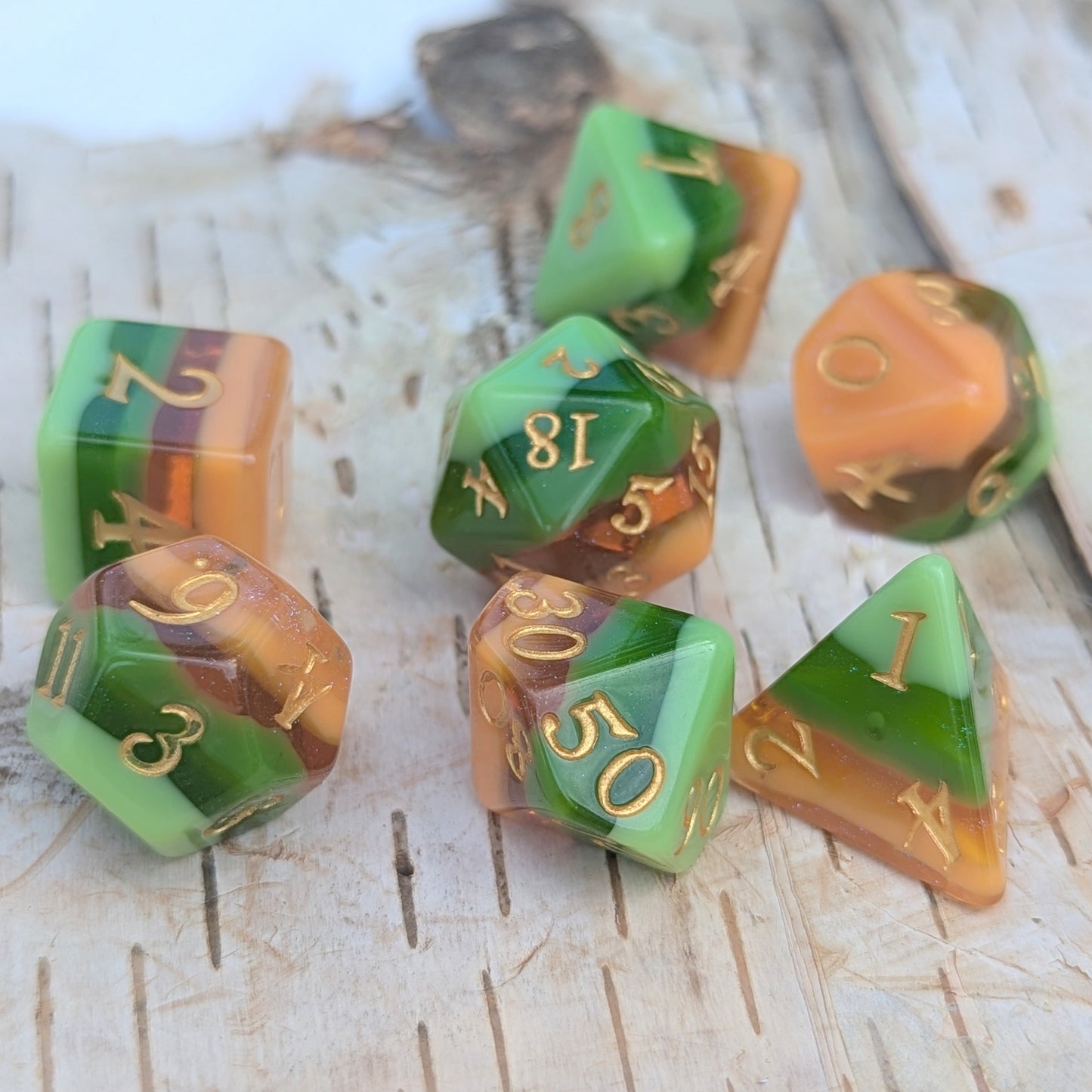 Timberlight Dice Set with green, brown, auburn, and yellow ombre layers, metallic gold numbers, placed on a wooden surface.