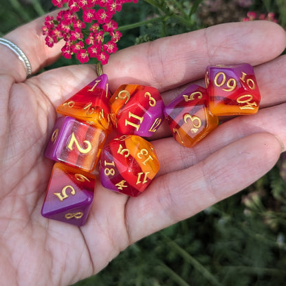 Vibrant Emberstorm Dice Set with fiery ombre colors from purple to orange, displayed on an open hand with flowers in the background.