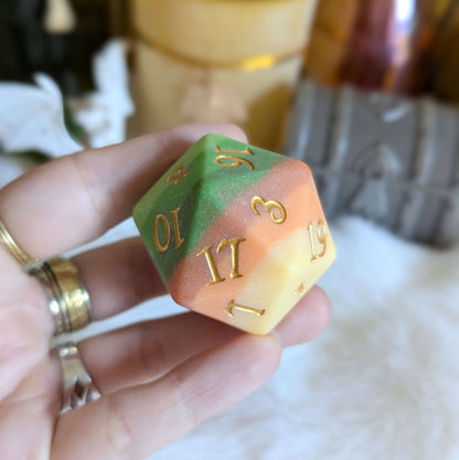 Hand holding Forest's Heart Large D20 with green, brown, and yellow segments and metallic gold numbers.