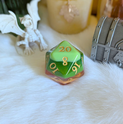 Green and gold D20 die on a fluffy white surface with dragon and treasure chest figures in the background.