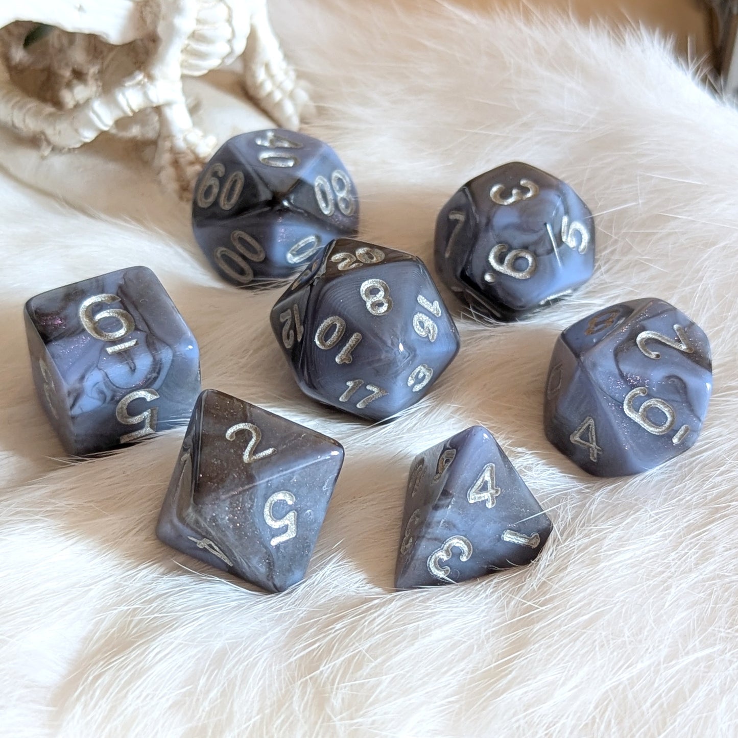 Moonlit Mirage round-edge dice set with grey-blue swirls, deep black streaks, and pink glitter accents on a soft surface.