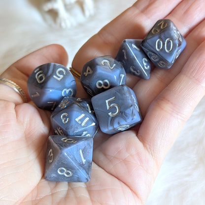 Hand holding Moonlit Mirage Dice Set with grey-blue marbled swirls and deep black streaks, featuring enchanting pink glitter accents.