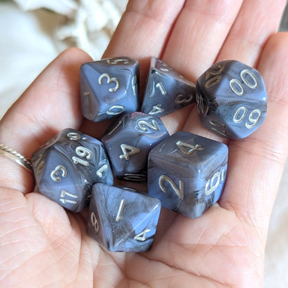 Moonlit Mirage round-edge dice set with grey-blue marbled swirls and silver numbers in a hand.