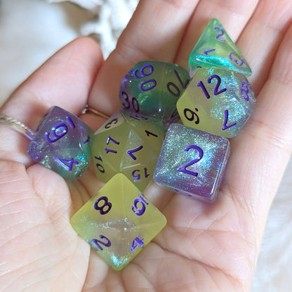 Dreamlight Grove glow-in-the-dark dice set with green-yellow, teal, and purple swirls and purple numbers, held in a hand.