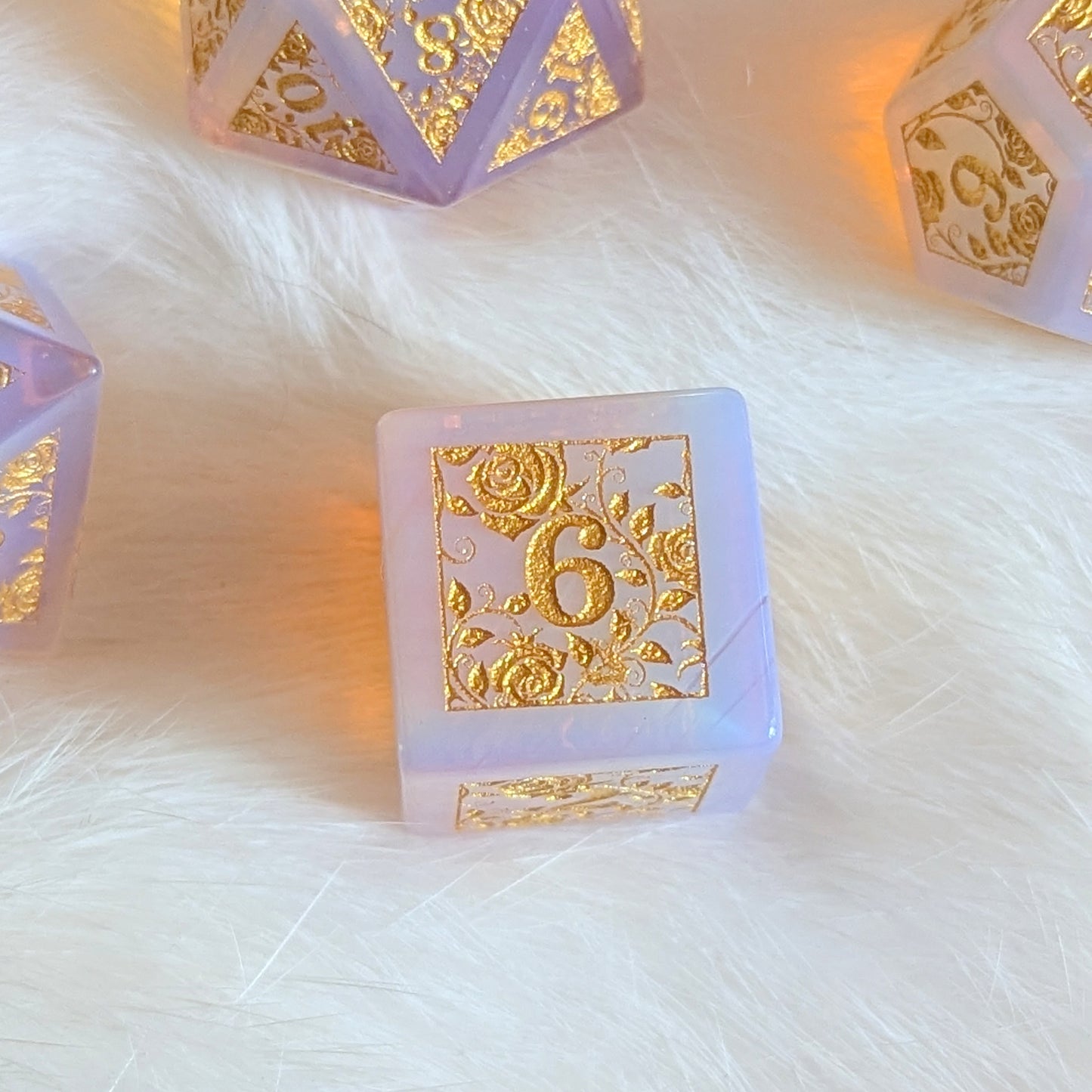 Pink opalite gemstone dice set with gold rose vine engravings on soft fur background.