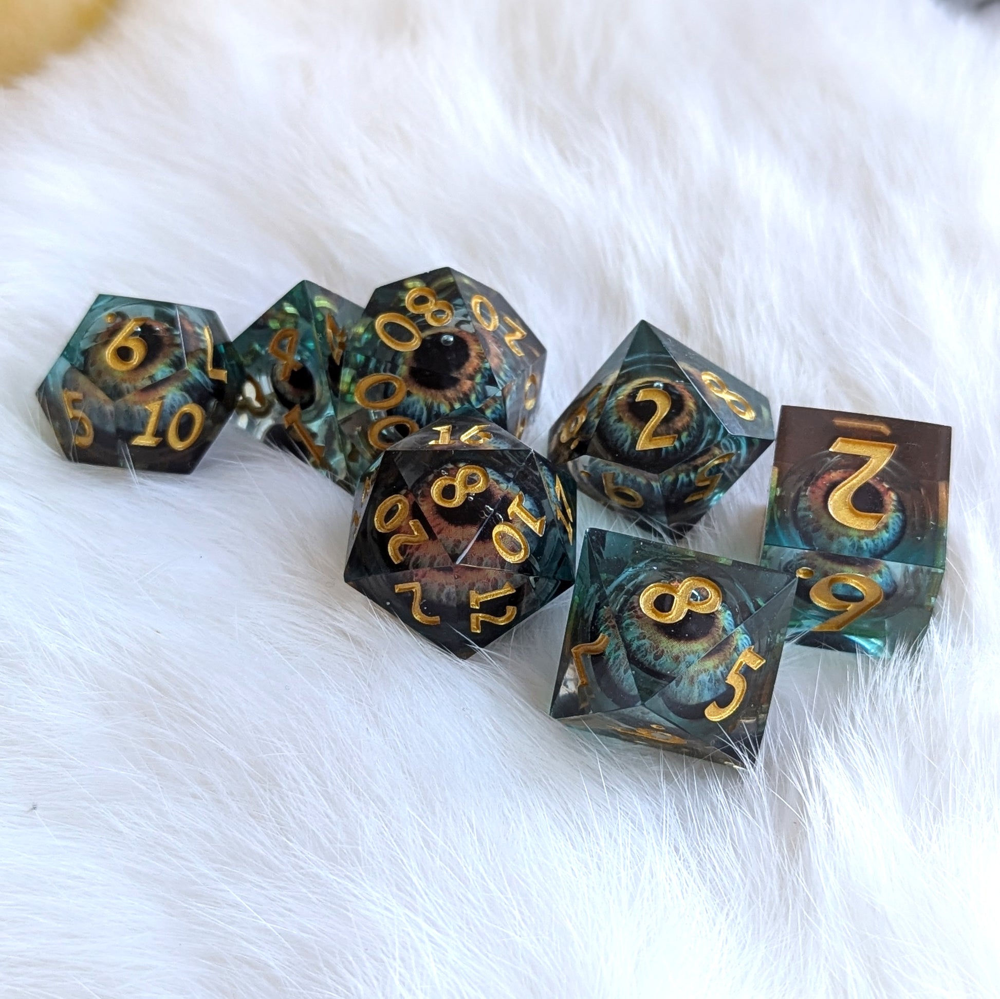 Watcher's Gaze Liquid Core Sharp Edge Dice Set featuring mystical eye design in blue and black resin with gold numbers on a white surface.