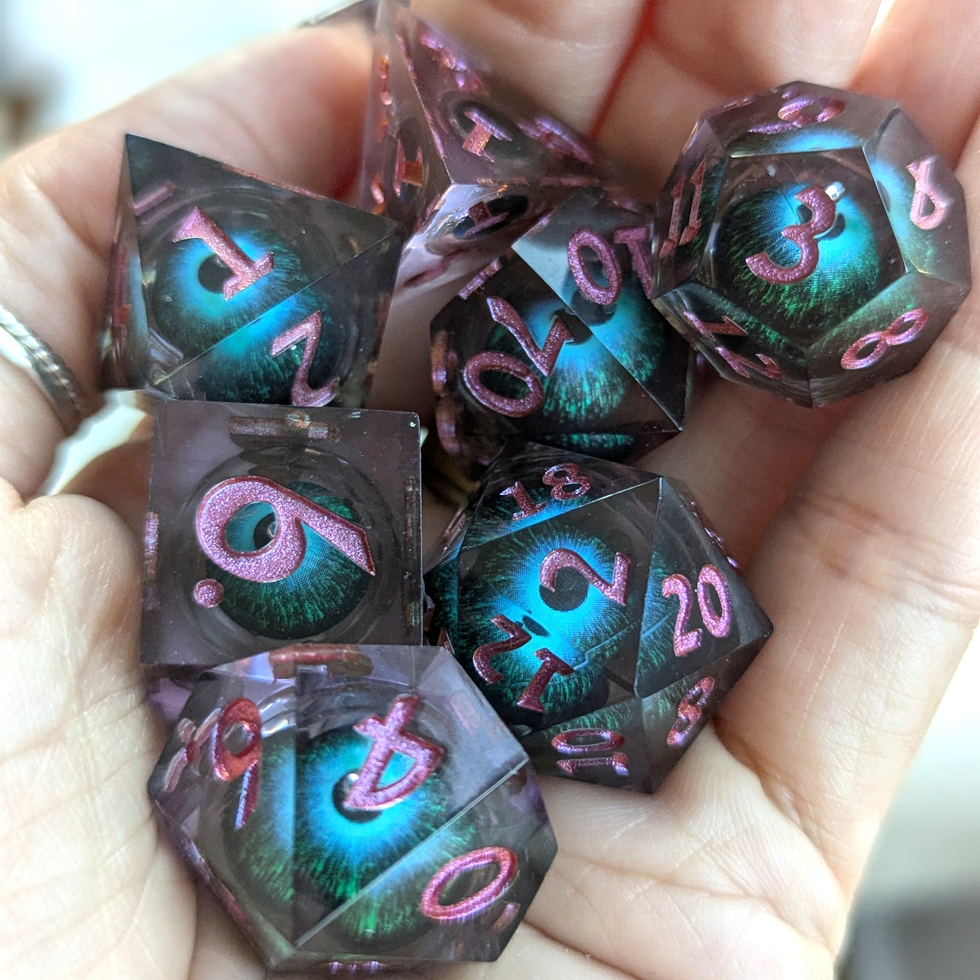 Hand holding Mystic Eye Liquid Core Sharp Edge Dice Set, featuring blue and green eye design in purple resin with pink numbers.