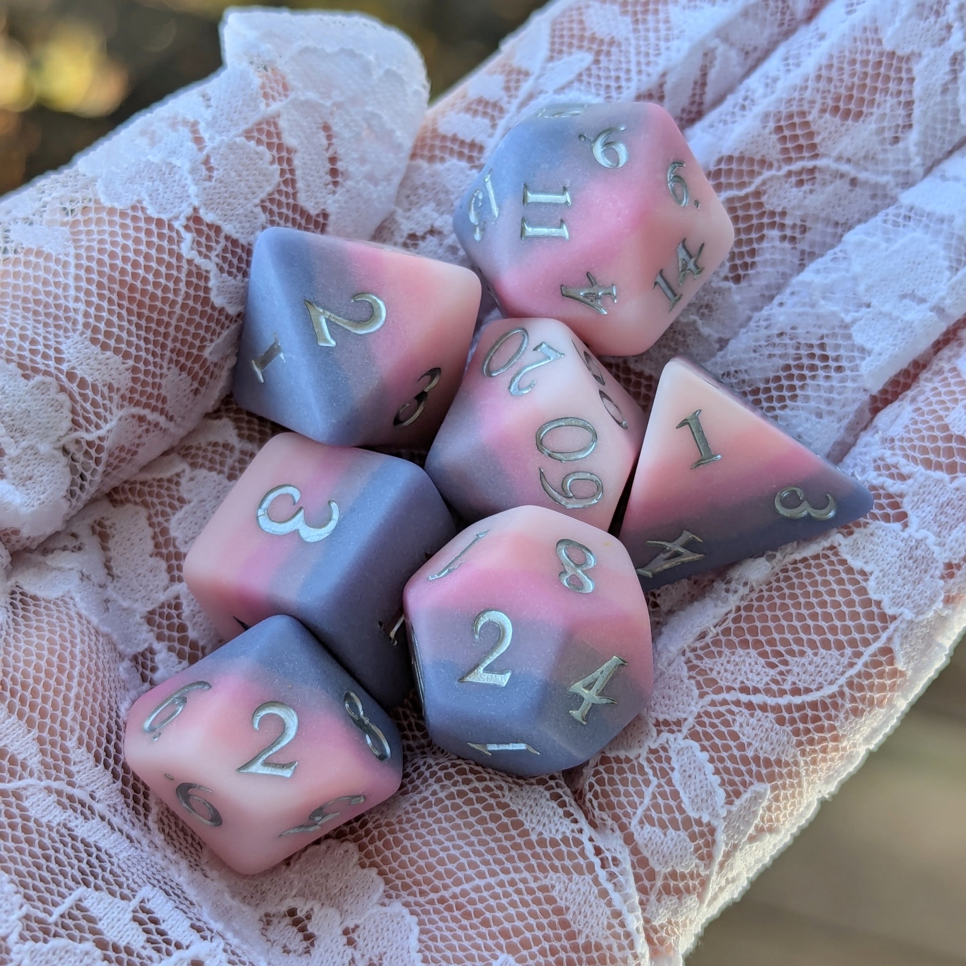 Dreamweaver Dice Set showcasing soft pink and lavender colors with silver numbers, held gently in a lace-clad hand.