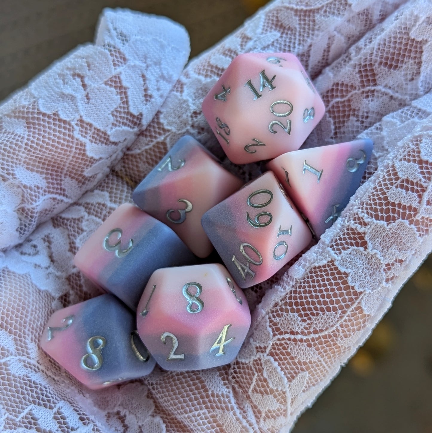 Dreamweaver Dice Set displayed in a hand with velvety ombre colors of pink and lavender, featuring silver-painted numbers.