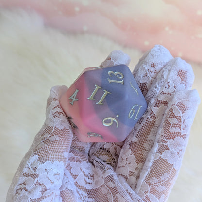 Dreamweaver Large D20 die in hand, featuring a matte ombre design of pinks and lavender with metallic numbers.