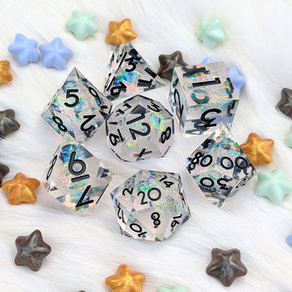 A set of holographic clear polyhedral dice with black printed numbers, arranged on a white faux fur surface with scattered pastel star-shaped candies.