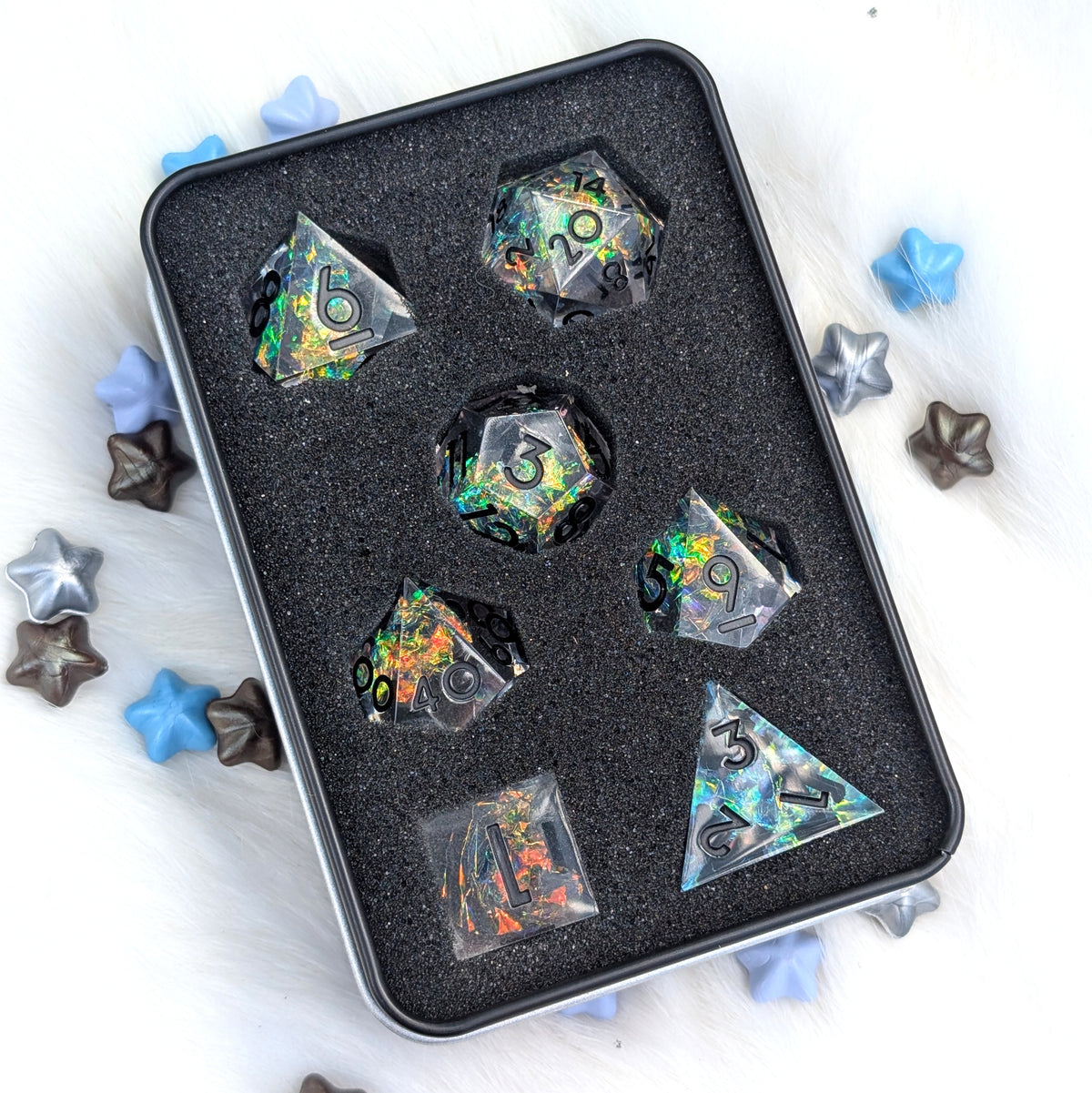 A set of holographic polyhedral dice displayed inside a black foam-lined metal tin, surrounded by scattered star-shaped decorative pieces on a white faux-fur surface.