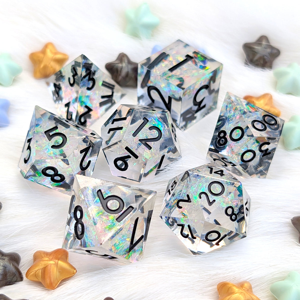 A set of holographic polyhedral dice with black printed numbers, scattered on a white faux fur surface with small pastel and metallic star-shaped props.
