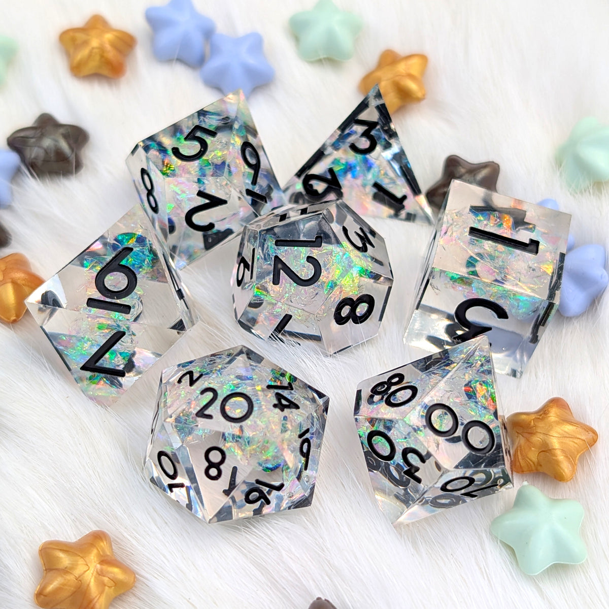 A set of holographic clear polyhedral dice with black printed numbers, arranged on a white faux fur surface with colorful star-shaped decorative props.