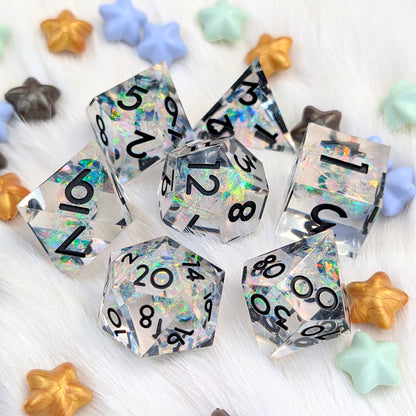 A set of holographic clear polyhedral dice with black printed numbers, arranged on a white faux fur surface with colorful star-shaped decorative props.