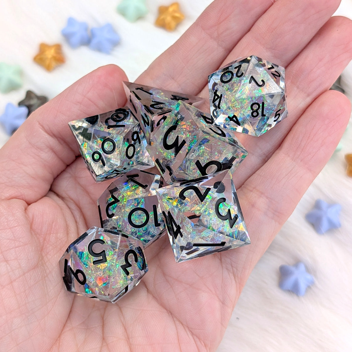A set of holographic clear polyhedral dice with black printed numbers, held in a person's hand. The background is a light surface with scattered pastel star-shaped beads.