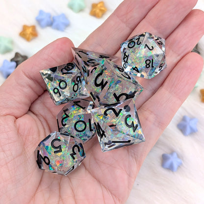 A set of holographic clear polyhedral dice with black printed numbers, held in a person's hand. The background is a light surface with scattered pastel star-shaped beads.