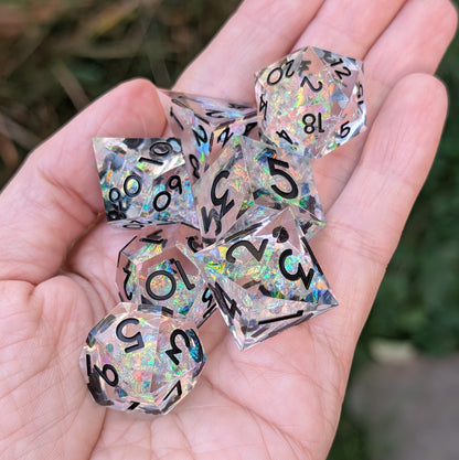 A set of holographic clear polyhedral dice being held in a person's hand, with a blurred outdoor background.