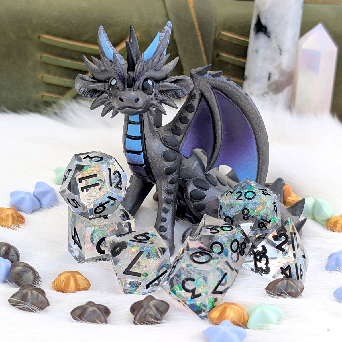 A fantasy dragon figurine in dark gray with blue accents sits surrounded by a matching set of holographic polyhedral dice on a white faux-fur surface, with crystals and shells in the background.