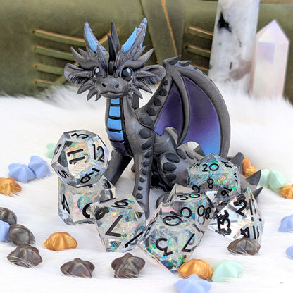 A fantasy dragon figurine in dark gray with blue accents sits surrounded by a matching set of holographic polyhedral dice on a white faux-fur surface, with crystals and shells in the background.