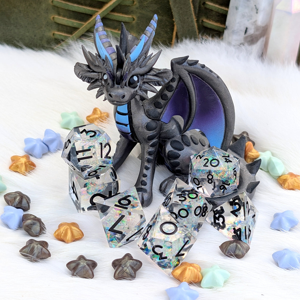 A fantasy dragon figurine in dark gray with blue horns and wing accents, posed among clear glittery polyhedral dice on a white faux-fur surface with small pastel and metallic star-shaped accessories.