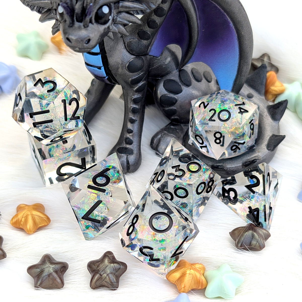 A set of holographic polyhedral dice with black printed numbers, arranged on a white textured surface with a dark dragon figurine and small star-shaped accessories.