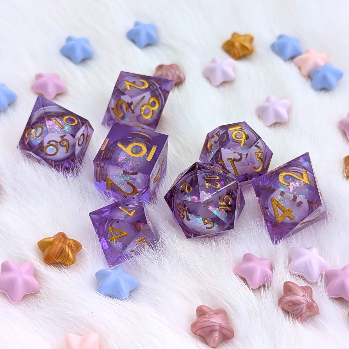 A set of translucent purple polyhedral gaming dice with gold numbers and iridescent glitter, arranged on a white faux-fur surface with pastel star-shaped beads.