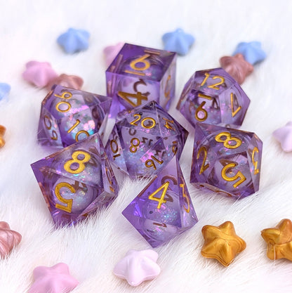 A set of translucent purple polyhedral gaming dice with gold numbers and glitter, arranged on a white faux fur surface with pastel star-shaped decorative props.