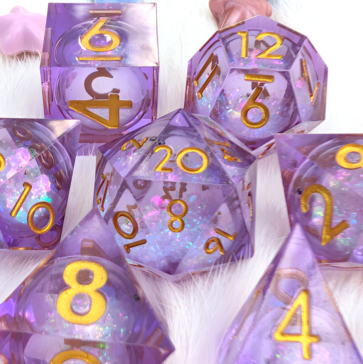 A set of translucent lavender polyhedral dice with gold-colored numbers and glittery internal details, arranged on a white faux fur surface.