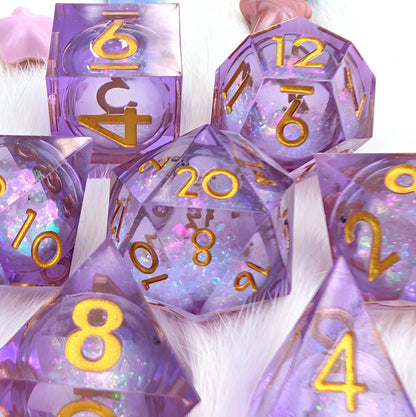 A set of translucent lavender polyhedral dice with gold-colored numbers and glittery internal details, arranged on a white faux fur surface.