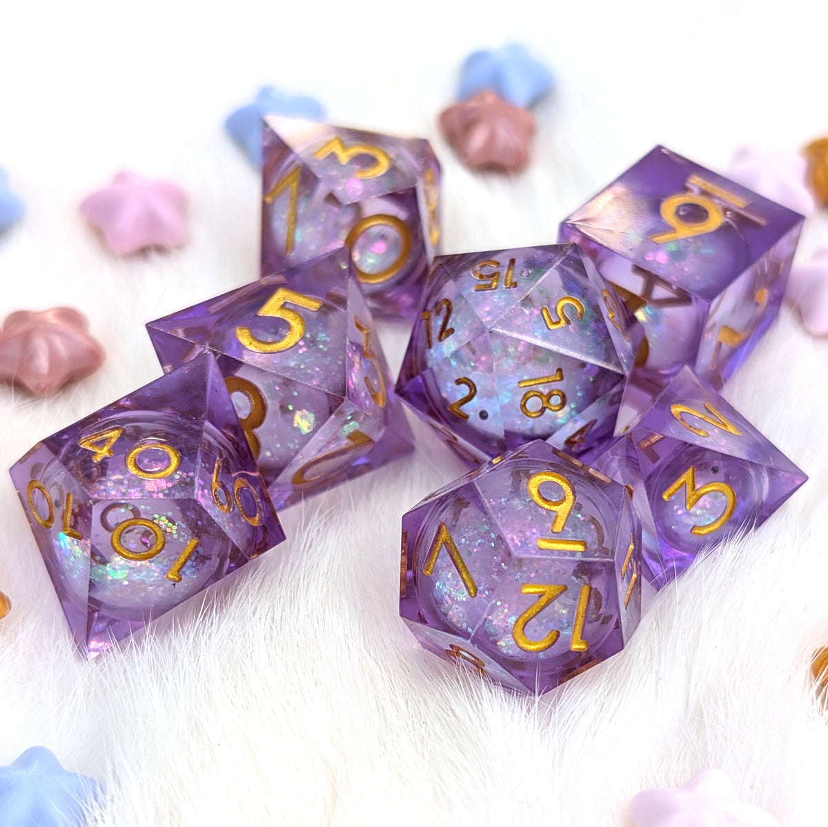 A set of translucent purple polyhedral gaming dice with gold numbering and iridescent glitter, arranged on a white furry surface with pastel star-shaped props.