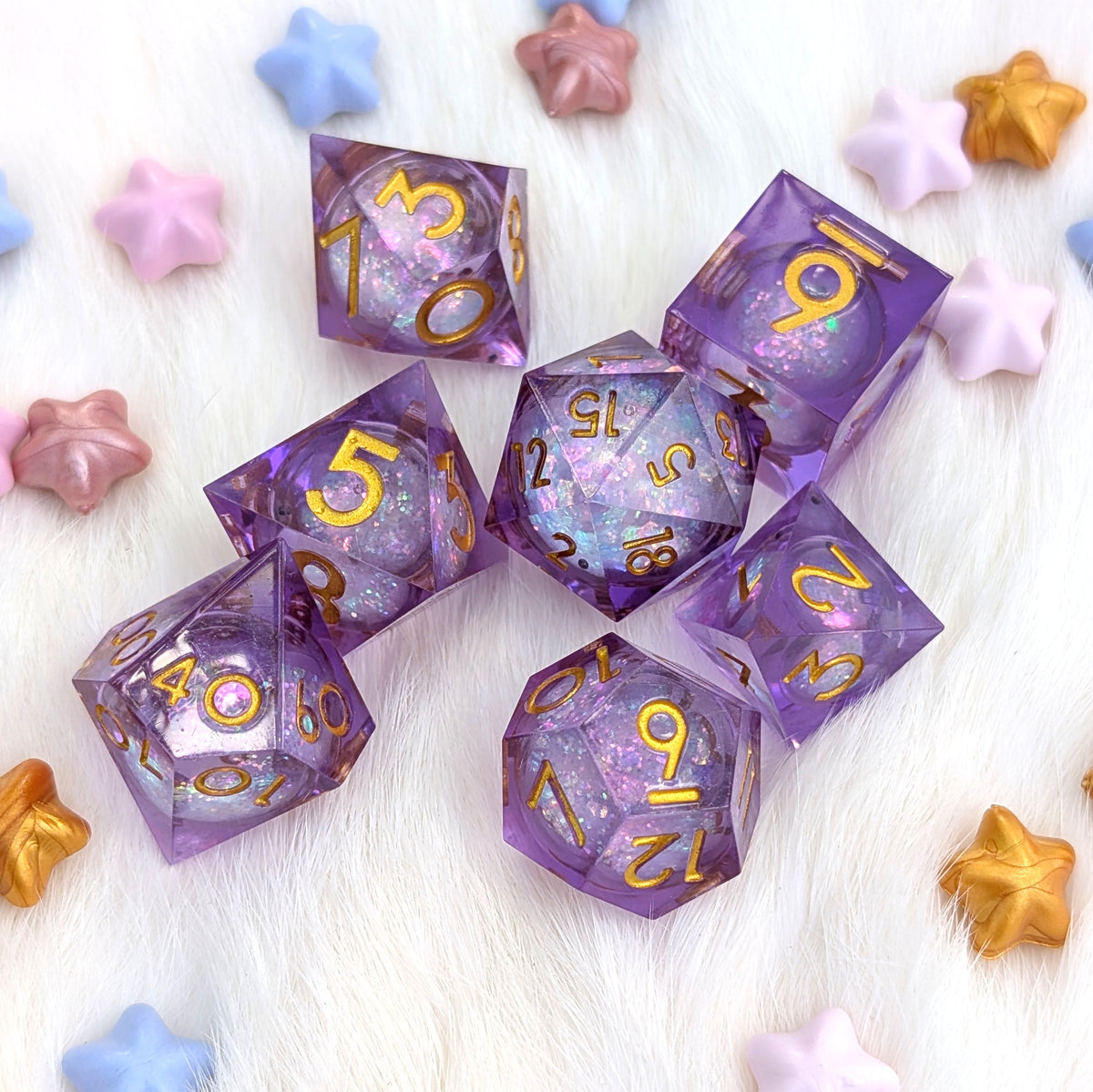 A set of purple glittery polyhedral gaming dice with gold numbers, arranged on a white faux fur background with pastel star-shaped props.