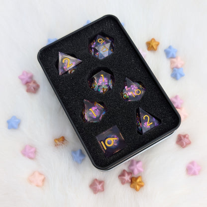 A set of translucent purple polyhedral gaming dice with gold numbers, displayed in a black foam tray inside a black metal box, surrounded by scattered pastel star-shaped candies on a white faux fur background.
