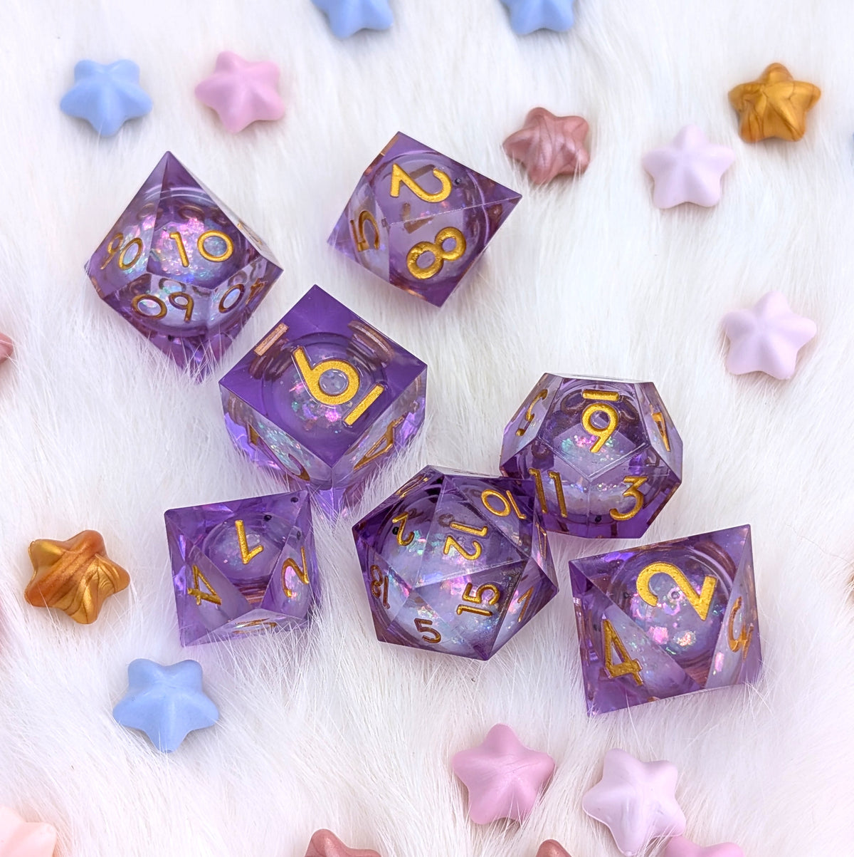 A set of purple translucent polyhedral dice with gold numbers and glittery pink sparkles, arranged on a white faux fur surface with pastel star-shaped decorative props.