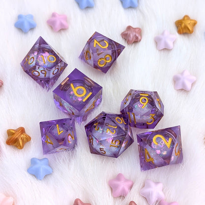 A set of purple translucent polyhedral dice with gold numbers and glittery pink sparkles, arranged on a white faux fur surface with pastel star-shaped decorative props.