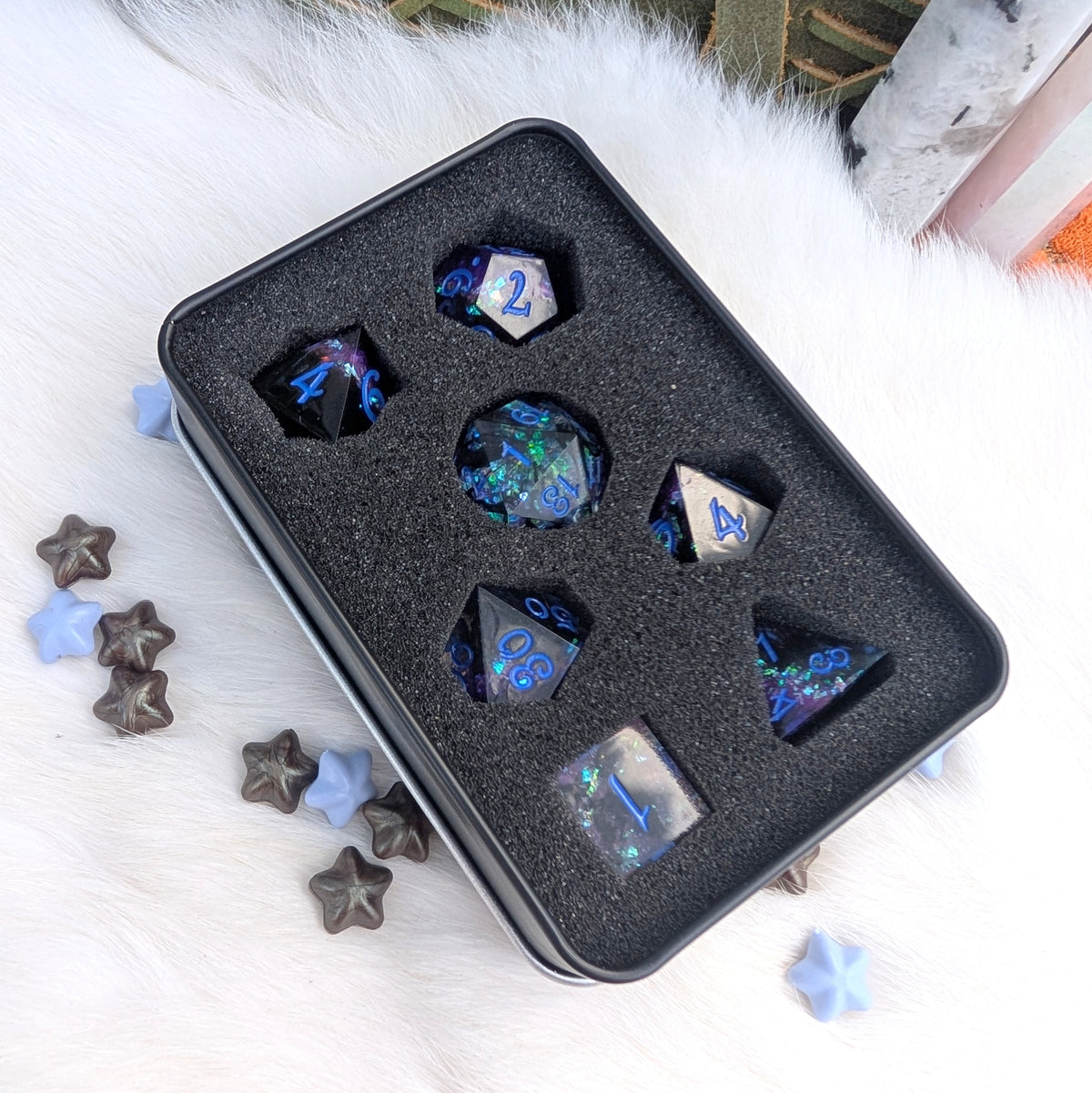 A set of polyhedral gaming dice displayed in a black foam-lined metal tin, photographed from above on a white faux-fur surface with decorative star-shaped pieces around it.