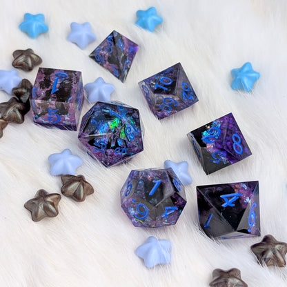 A set of translucent polyhedral dice with a purple and black galaxy pattern and glowing blue numbers, arranged on a white faux fur surface with decorative star-shaped props.