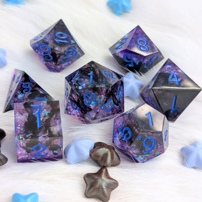 A set of purple and black glitter polyhedral gaming dice with blue numbers, arranged on a white faux fur surface with small decorative star and heart props.